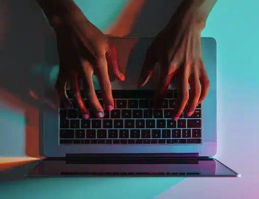 Close up photo of hands typing on a laptop with multi-colored room lighting